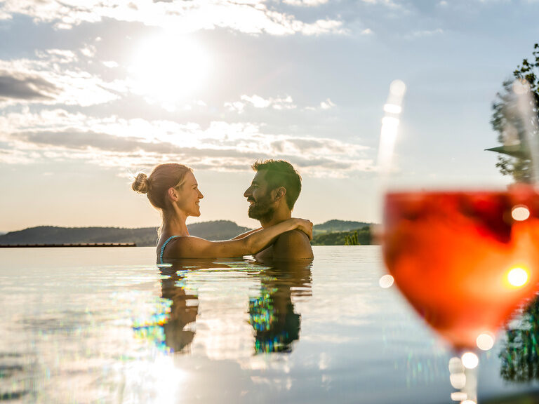 Pärchen umarmt sich bei Sonnenuntergang im Außenpool, Man sieht an der Seite noch einen frisch angerichteten Cocktail