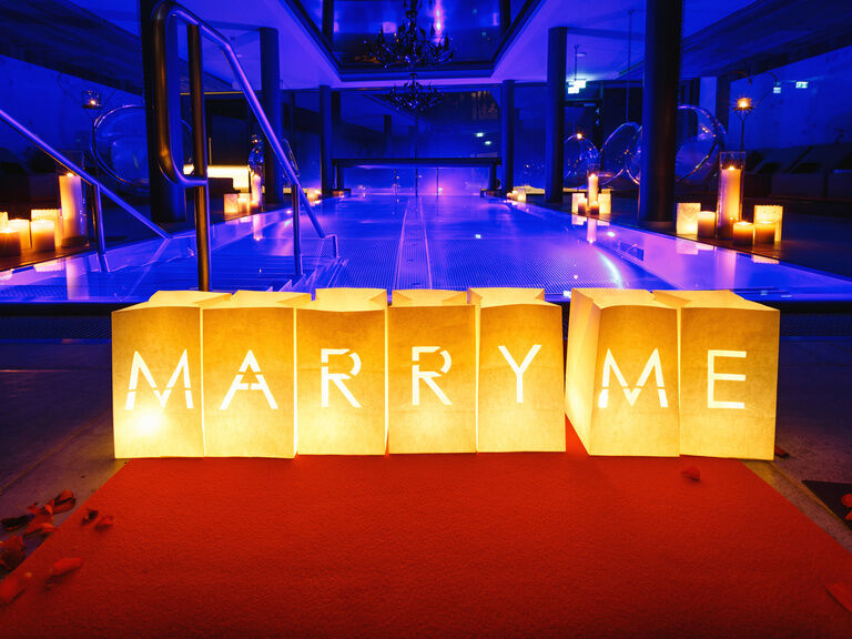 Papierboxen mit leuchtender Aufschrift 'Marry Me', im Hintergrund beleuchteter Innenpool