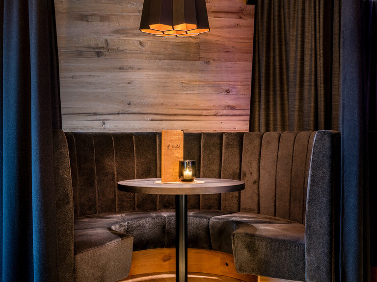 Runder Tisch umrahmt von Sitzbank und verstellbaren Vorhängen im Luxushotel Hüttenhof Runder Tisch mit Bodenbeleuchtung umrandet von grauer Sitzbank und verstellbarem Vorhang