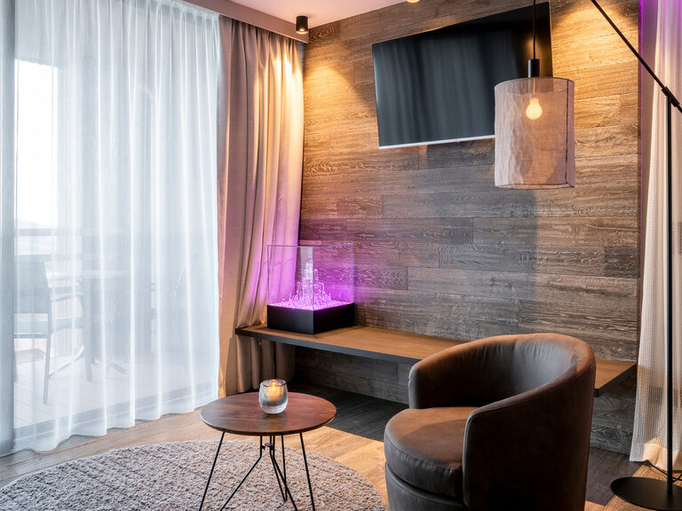 Sessel mit Beistelltisch, mit Blick auf Balkon, dahinter Holzwand mit montiertem Flachbild-TV