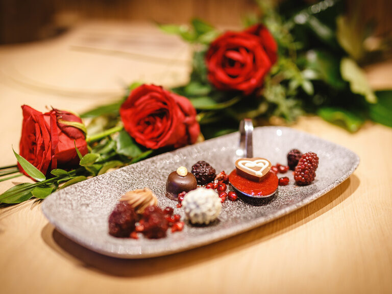 Dessert mit Früchten und feinen Pralinen, auf einem rechteckigem Teller, daneben ein Rosenstrauß